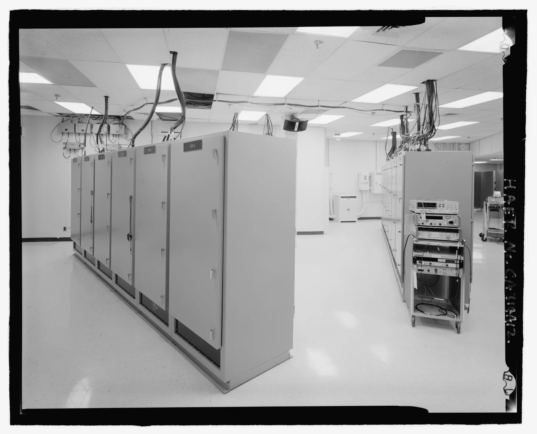 Black-and-white photograph of a computer maintenance operations center: a row of labelled equipment cabinets with cable bundles running overhead, and a wheeled test cart alongside