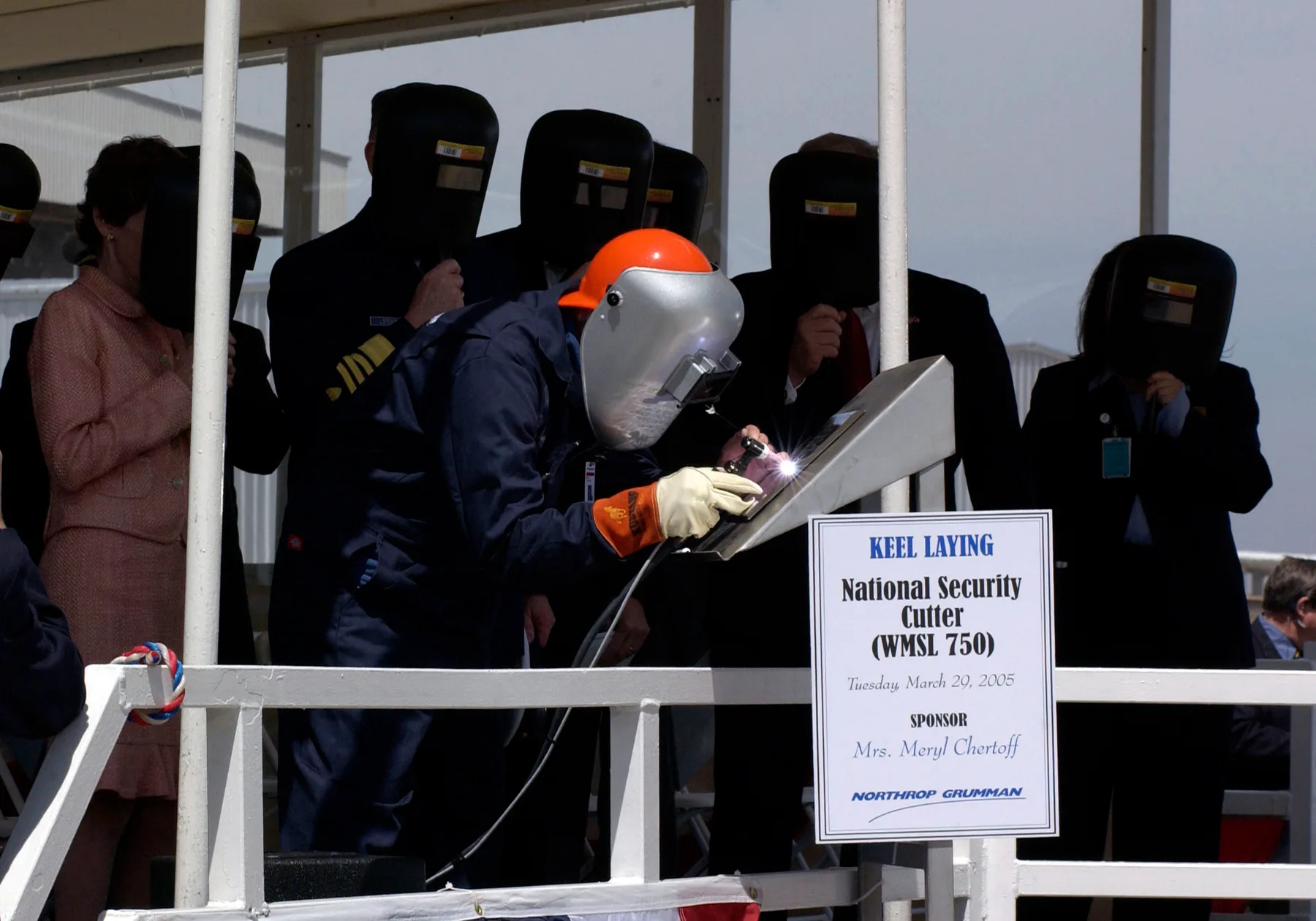 A welder in a hard hat and mask strikes an arc on a placard reading Keel Laying, National Security Cutter WMSL 750, while officials in welding masks look on